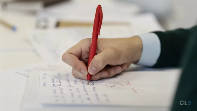 Mão de uma pessoa segurando uma caneta vermelha e corrigindo um texto manuscrito em folha pautada, com marcações em vermelho