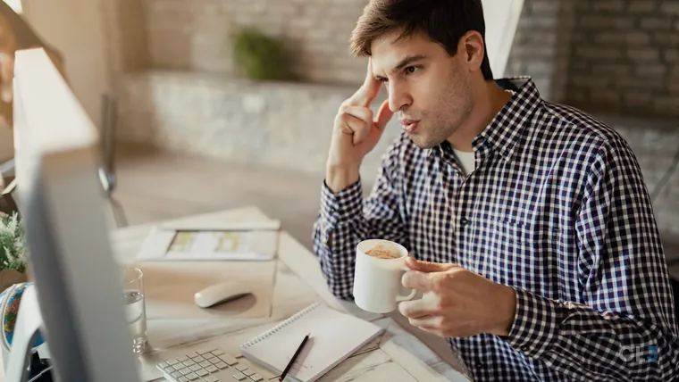 Jovem empreendedor com expressão preocupada diante do computador, segurando uma xícara de café no escritório