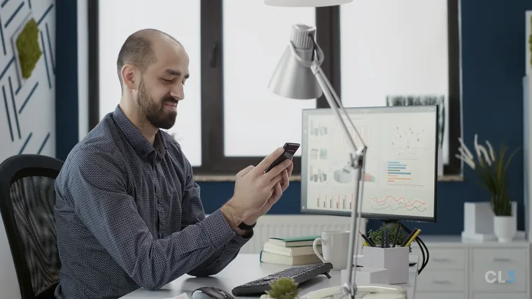 Profissional analisando estatísticas de campanha no celular, com o computador ao fundo no escritório